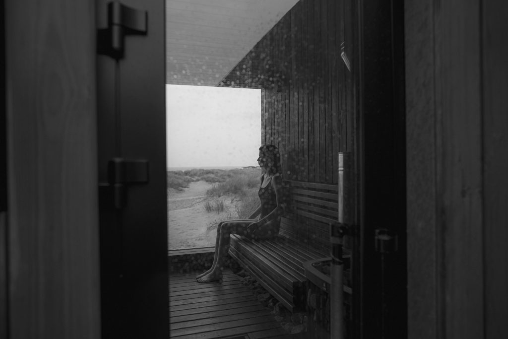 A woman sits in a sauna gazing out at a tranquil landscape through a glass door.