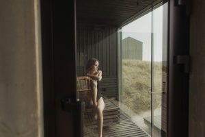 A woman in a bikini relaxes inside a cozy wooden sauna with a view of grassy dunes outside.