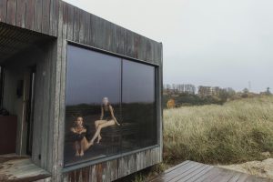 Two women in swimsuits relaxing in a wooden sauna with glass window overlooking nature.